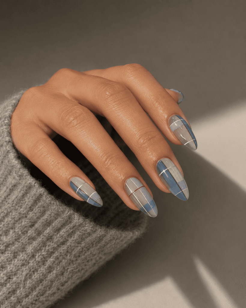 Soft blue plaid nail art on almond-shaped nails with delicate white line details, styled with a cozy gray sweater in warm natural lighting. Trendy minimalist manicure inspiration perfect for winter nails, classy acrylic nail designs, and cute nail inspo.
