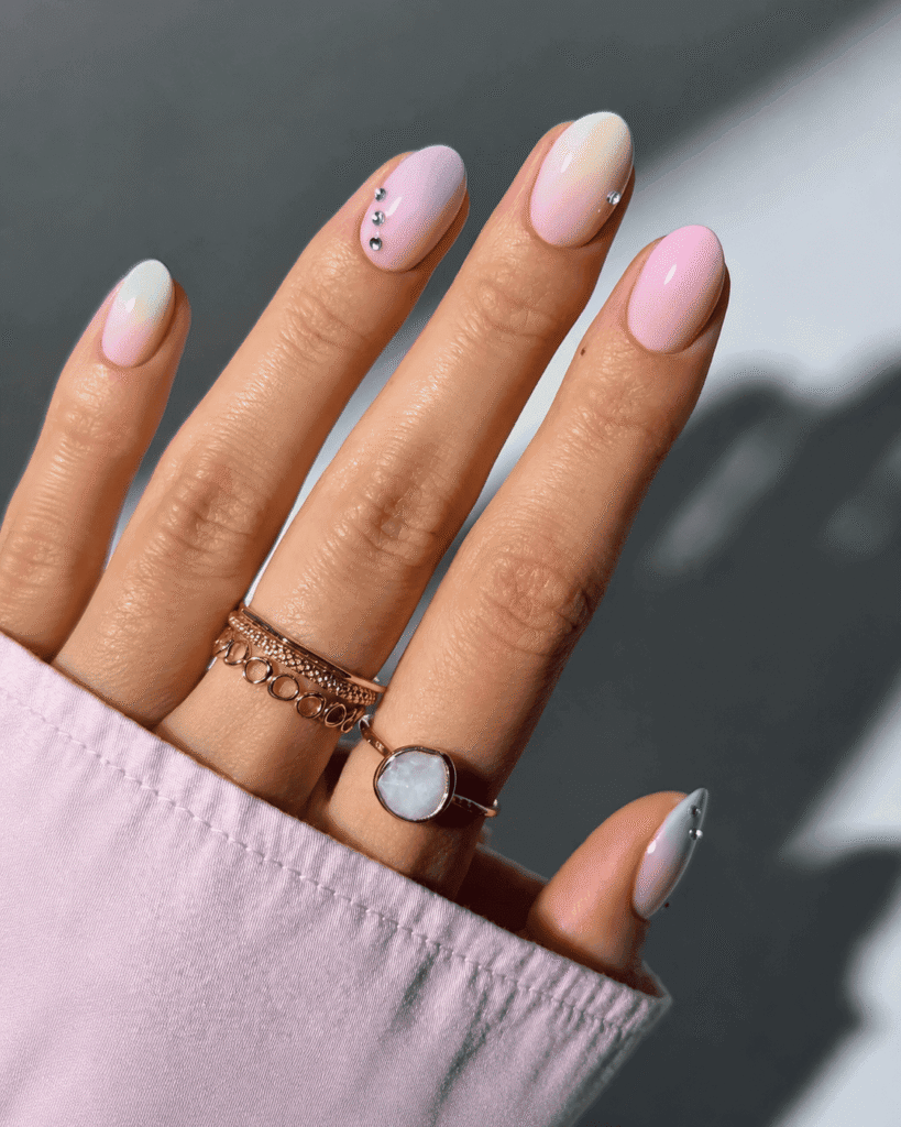 Soft pastel pink and white almond-shaped nails with glossy finish, delicate rhinestone accents, and elegant minimalist nail art on a hand wearing rose gold rings and a light pink sleeve.