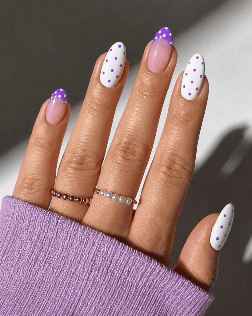 Cute lavender and white polka dot nail art with glossy almond-shaped nails, featuring pastel purple French tips and playful dotted designs, styled with delicate rings and a cozy lilac sweater sleeve.