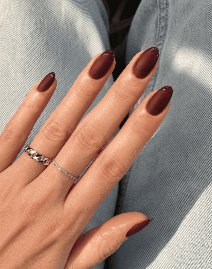 Close-up of elegant burgundy ombré nails with a soft matte finish and glossy dark red French tips, styled on almond-shaped nails. The hand is resting on light denim jeans and accessorized with silver rings, creating a classy and trendy luxury nail art look perfect for fall and winter nail inspiration.
