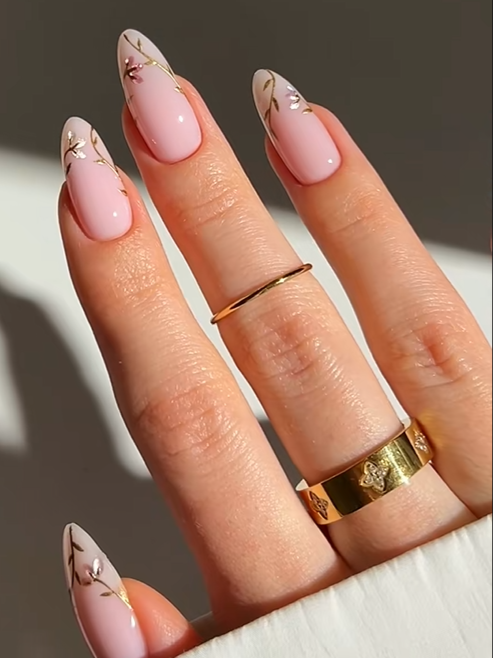Elegant almond nails with glossy soft pink polish and delicate gold floral branch nail art, creating a classy and feminine manicure aesthetic.