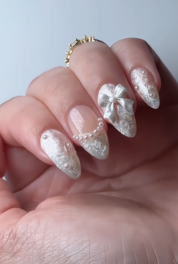 Elegant white pearl almond nails with 3D floral texture, pearl accents, and a decorative bow design, creating a luxurious bridal-inspired manicure aesthetic.